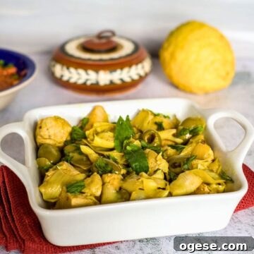 Moroccan-Spiced Keto Chicken with Harissa and Olives 4 A white ceramic dish contains a cooked Keto Moroccan chicken meal with cubed chicken, artichokes, olives, and herbs. In the background, there is a round bread and a painted container.