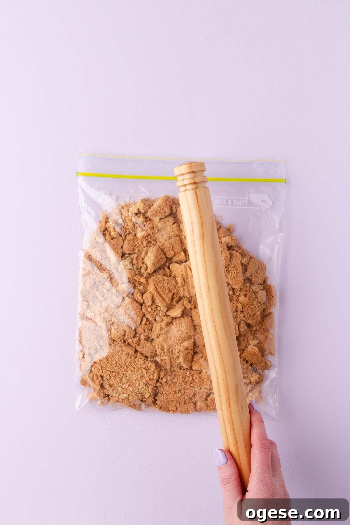 Plain biscuits in a resealable plastic bag, being crushed into small pieces with a wooden rolling pin.