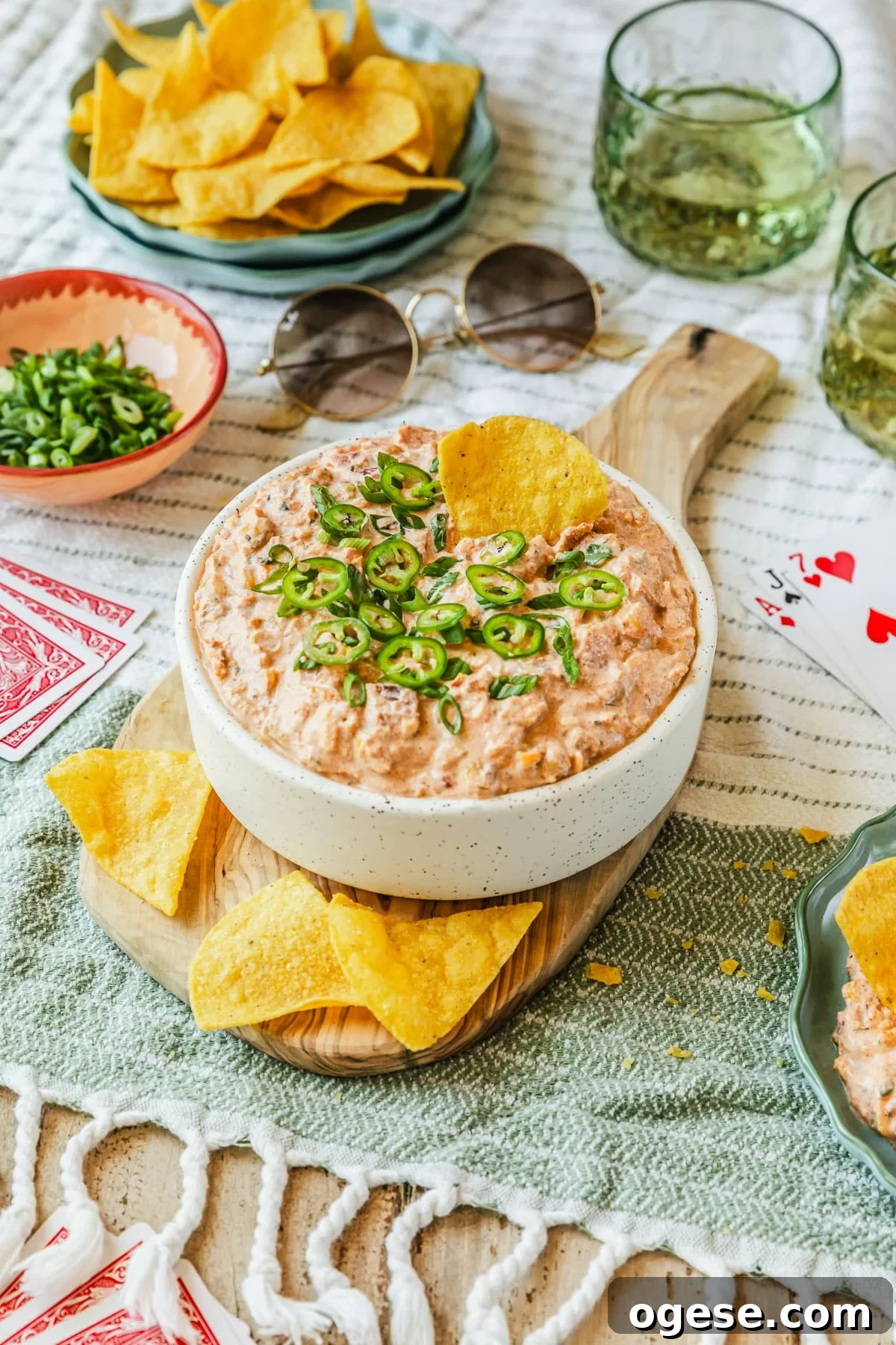 A white bowl of boat dip with corn on a wood board next to glasses of beer, a pink bowl of green onions, a blue bowl of corn chips, cards, and sunglasses with a white and green striped background.