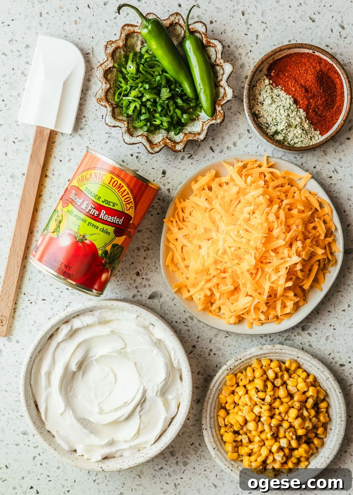 Brown and white bowls of cheddar cheese, sour cream, corn, ranch seasoning, taco seasoning, and green onions next to a can of tomatoes on a white terrazzo counter.
