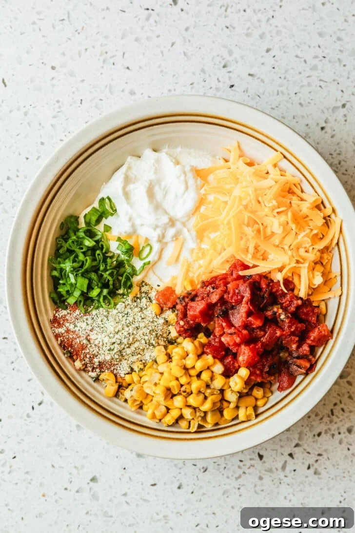 A white bowl of sour cream, seasonings, tomatoes, cheddar cheese, corn, and green onions on a white terrazzo counter.