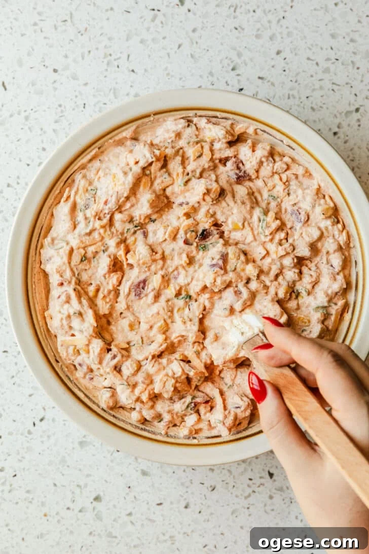 A hand using a rubber spatula to mix boat dip together in a white bowl.
