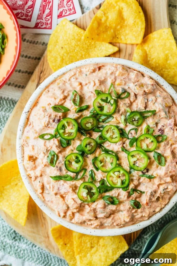 A white bowl of boat dip on a wood board of tortilla chips next to cards and a pink bowl of scallions on a green and white striped blanket.