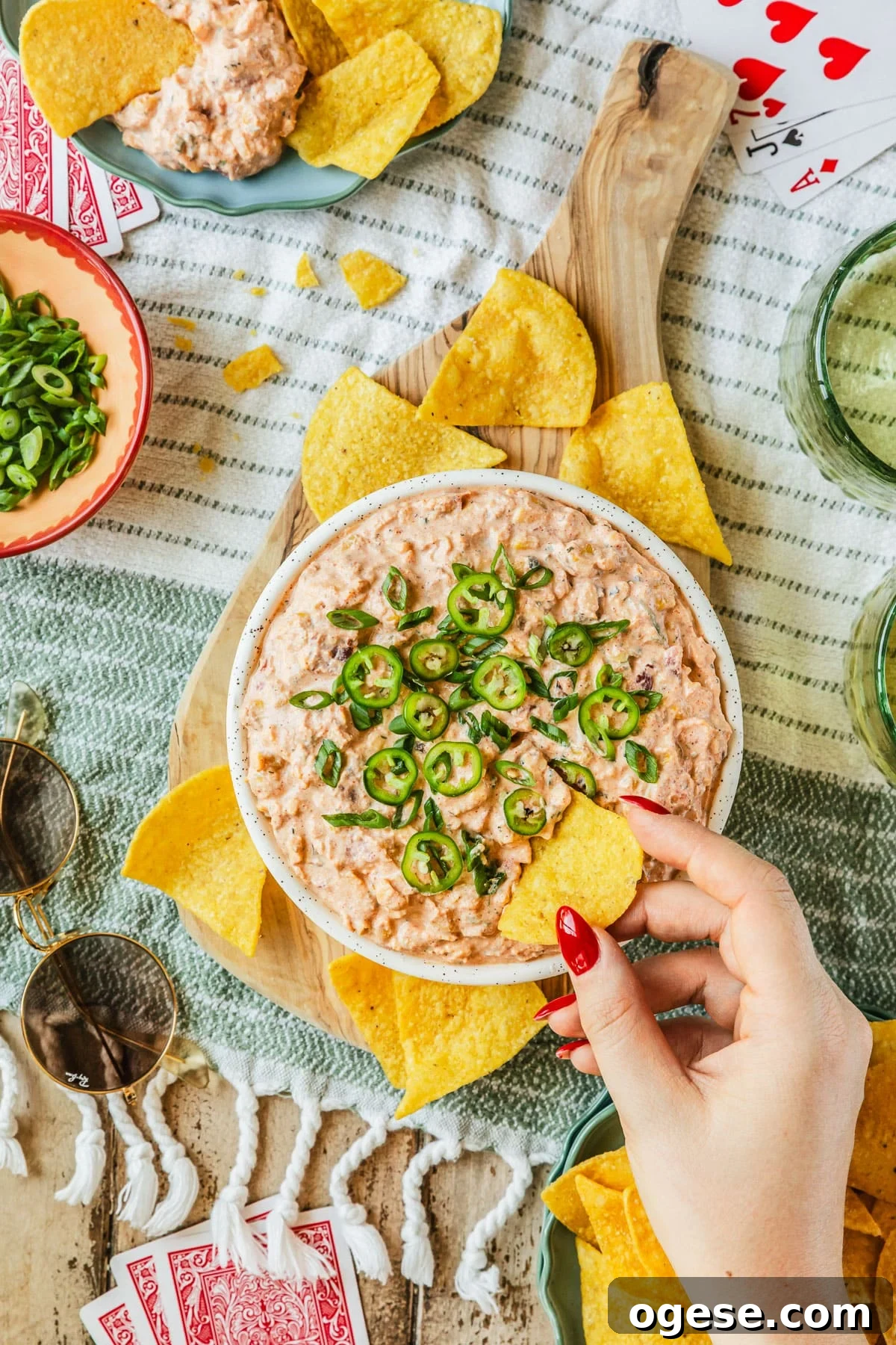 A woman's hand using a corn chip to scoop boat dip out of a white bowl on a striped picnic blanket next to glasses of beer, playing cards, sunglasses, and a blue bowl of corn chips.