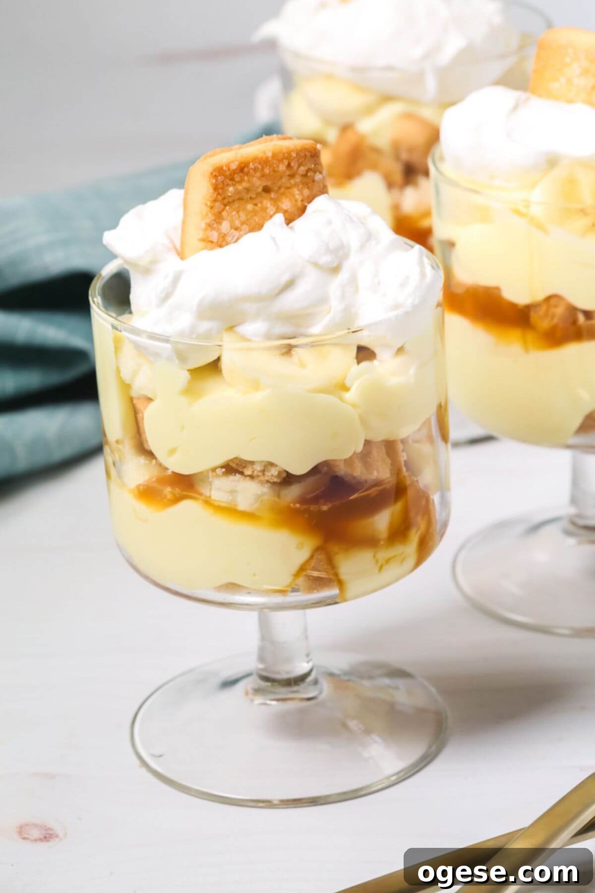 An easy banana pudding delight: a glass dessert cup layered with creamy pudding, banana slices, caramel sauce, and fluffy whipped cream, all crowned with a cookie. Two similarly tempting cups linger in the background.