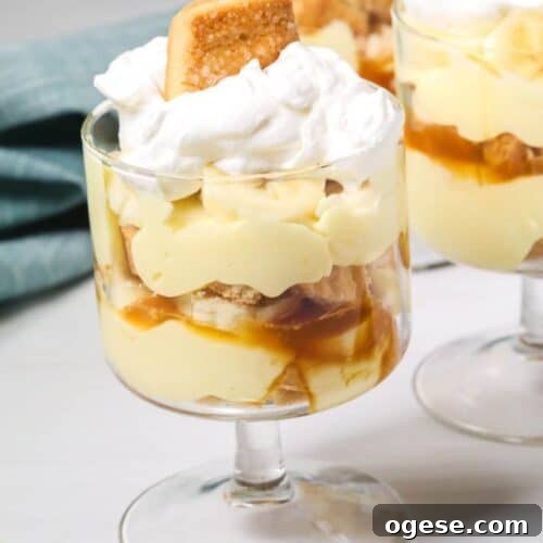 An easy banana pudding delight: a glass dessert cup layered with creamy pudding, banana slices, caramel sauce, and fluffy whipped cream, all crowned with a cookie. Two similarly tempting cups linger in the background.