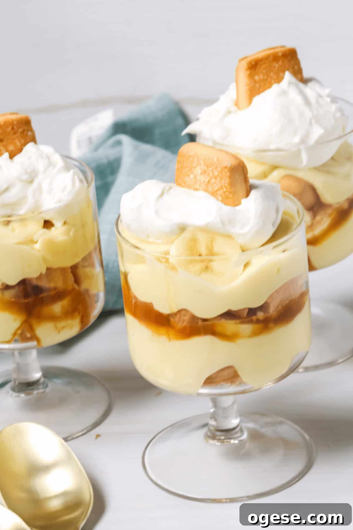 Three glass dishes of layered, dessert topped with whipped cream and a cookie, next to a blue napkin and a gold spoon.