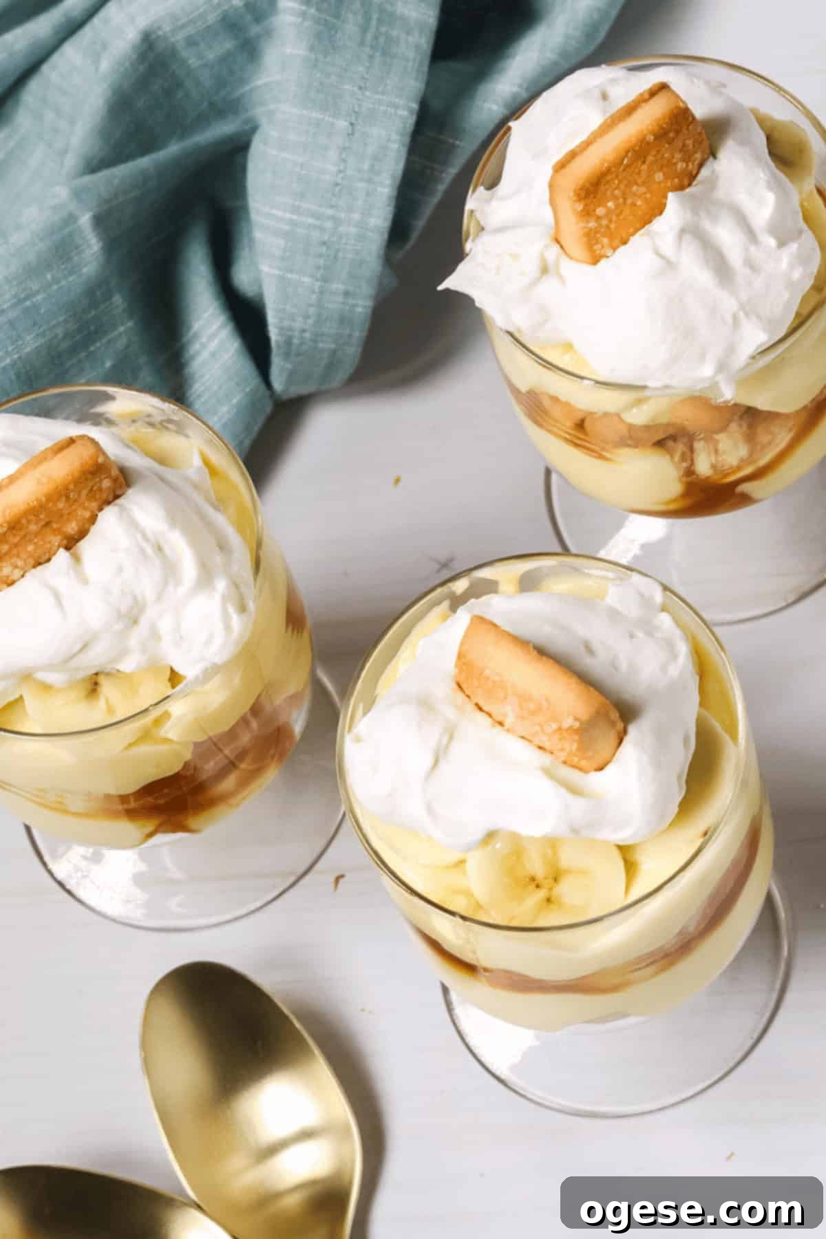 Three glasses of easy banana pudding topped with whipped cream and a cookie rest on a white surface. A blue cloth drapes beside them, along with two gold spoons, ready to scoop up the deliciousness.