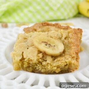 A close-up of a Peanut Butter Banana Bar topped with a banana chip, served on a white decorative plate.