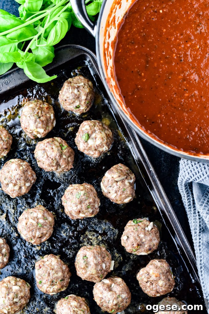 Authentic Homemade Pasta Sauce Recipe for Rich, Simple Flavor 7 Cooked meatballs on a baking sheet with a pan of spaghetti sauce and fresh basil leaves next to it.