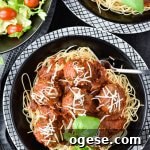 A black bowl of homemade spaghetti and meatballs on a white checkered plate. A salad and another bowl of spaghetti and meatballs sit in the corner of the photo.