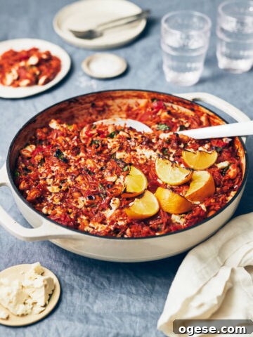 A white skillet filled with tomato lemon orzo on a table with smoked paprika, feta, and water glass in the background.