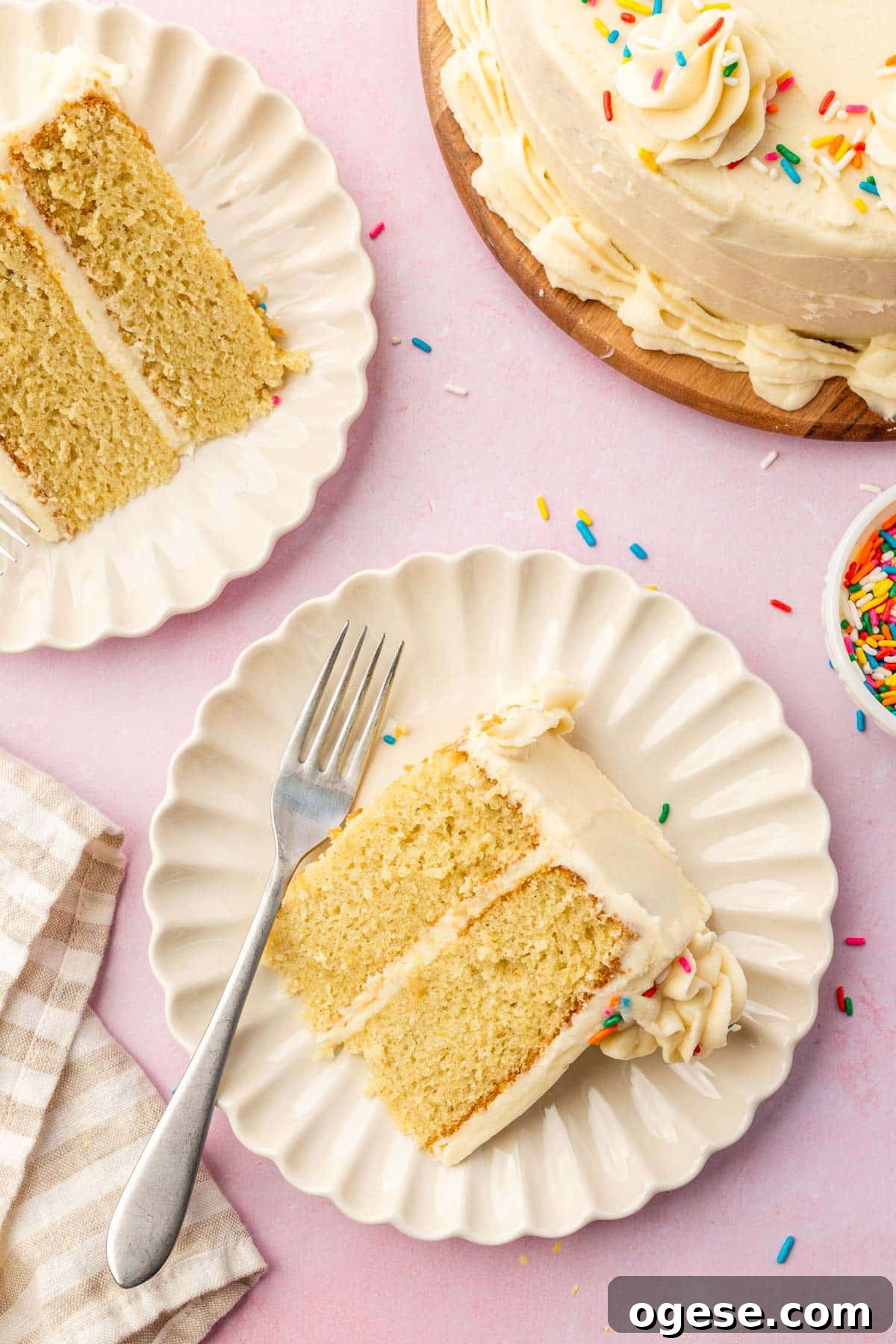 An overhead view of two dessert plates each with a single slice of gluten free vanilla cake with the larger cake peaking in to the photo.