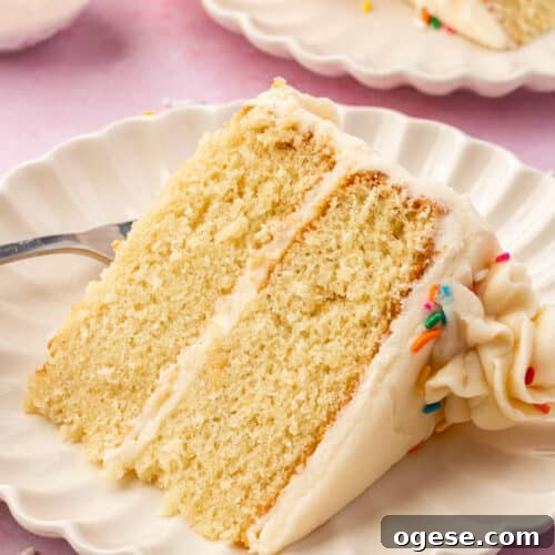 A slice of 2-layer gf vanilla cake with vanilla frosting and rainbow sprinkles on a dessert plate with another slice of cake and a bowl of rainbow sprinkles in the background.
