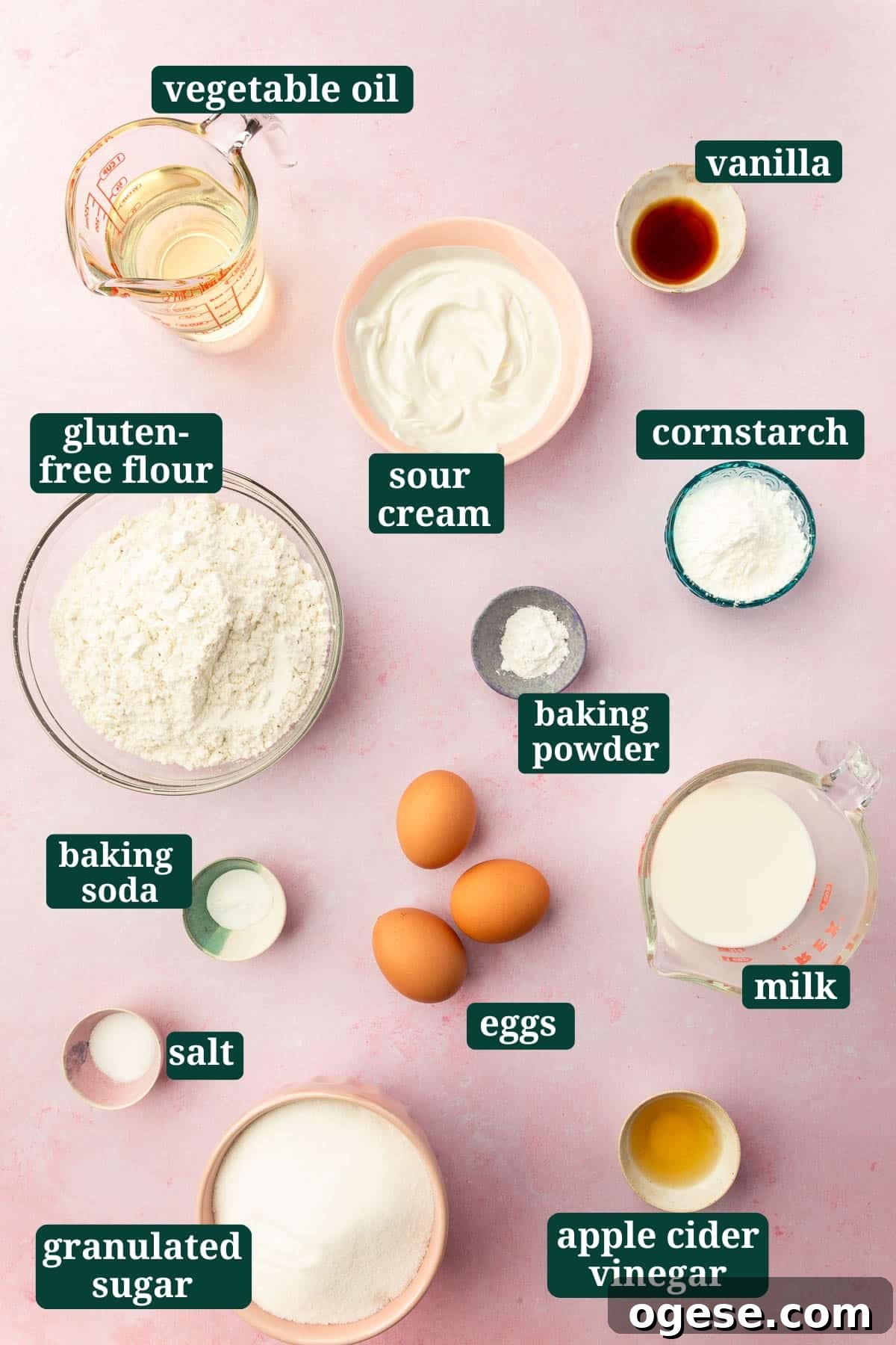 Ingredients in small bowls to make gluten-free vanilla cake including oil, sour cream, vanilla, gluten-free flour, baking powder, cornstarch, eggs, salt, baking soda, granulated sugar, apple cider vinegar and milk with text overlays over each ingredient.