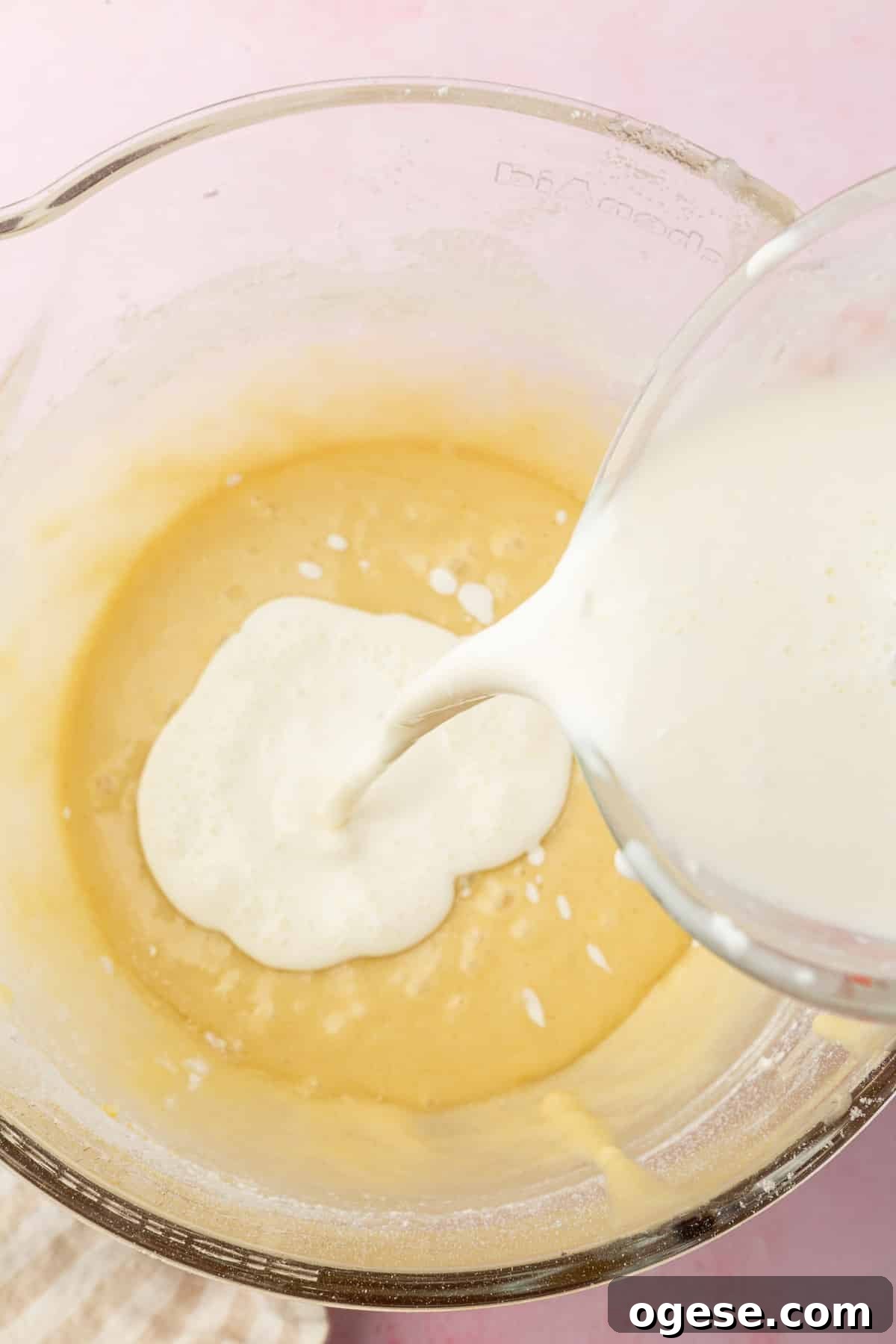 A glass measuring cup pouring a sour cream and milk mixture into a mixing bowl of gluten free cake batter.