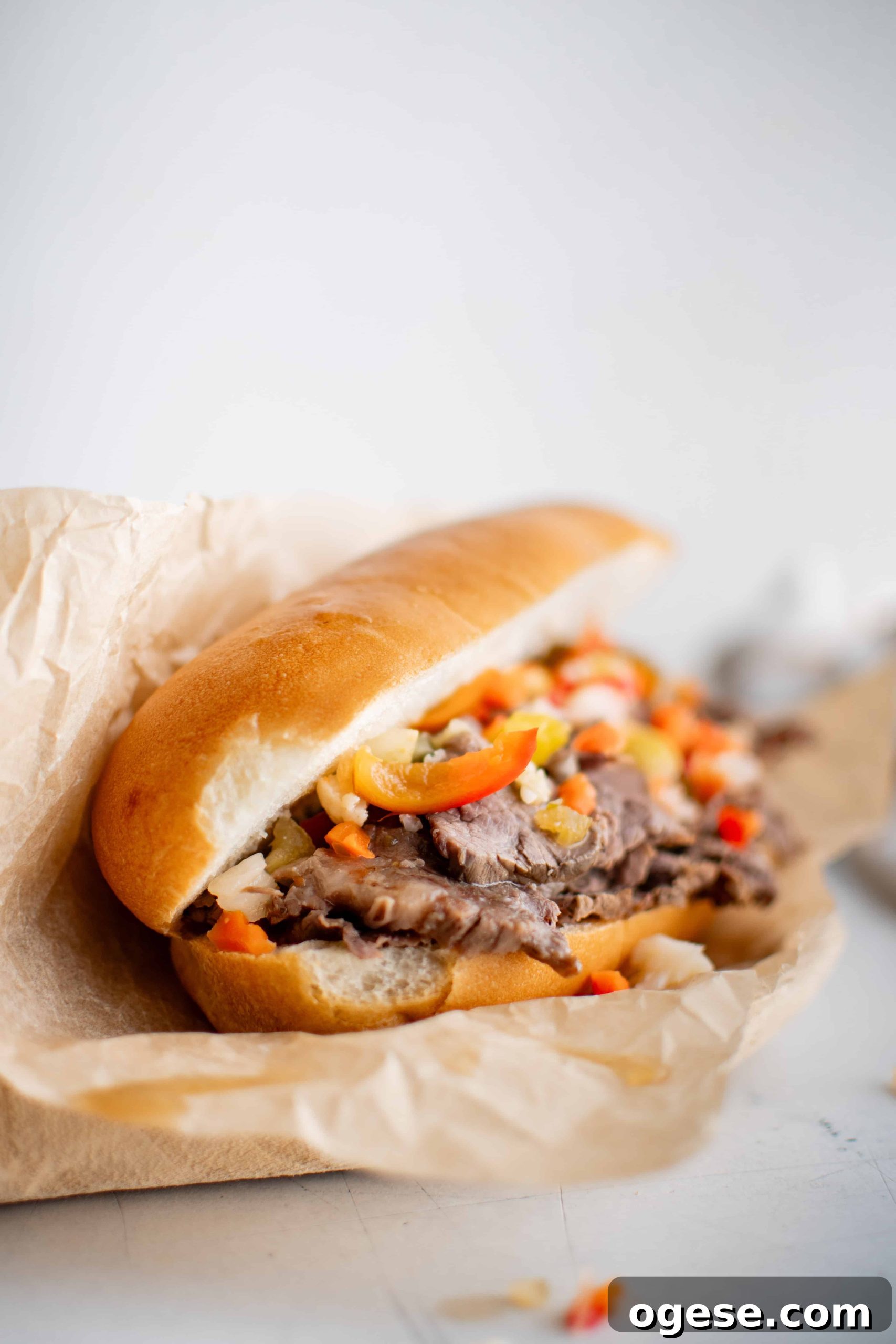 Classic Italian Beef Sandwich Recipe: Tender Slow-Cooked Roast Beef on Italian Bread 2 Italian roll topped with thinly sliced beef topped with chopped Giardiniera and hot cherry peppers served on a white plate lined with parchment paper.