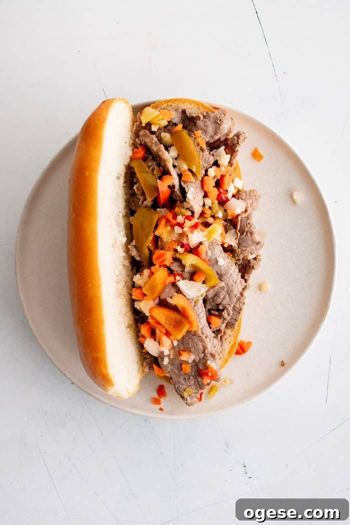 Classic Italian Beef Sandwich Recipe: Tender Slow-Cooked Roast Beef on Italian Bread 14 Italian roll with thinly sliced beef topped with chopped Giardiniera and hot cherry peppers served on a white plate.