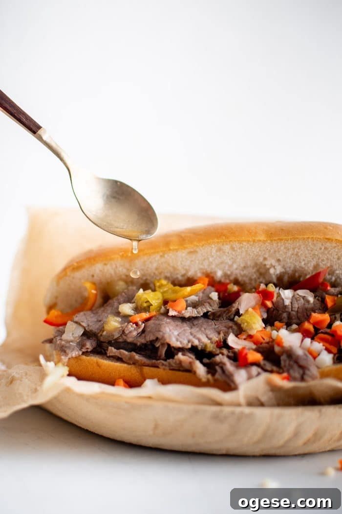 Classic Italian Beef Sandwich Recipe: Tender Slow-Cooked Roast Beef on Italian Bread 16 Drizzling au jus over an Italian roll topped with thinly sliced beef topped with chopped Giardiniera and hot cherry peppers served on a white plate lined with parchment paper.