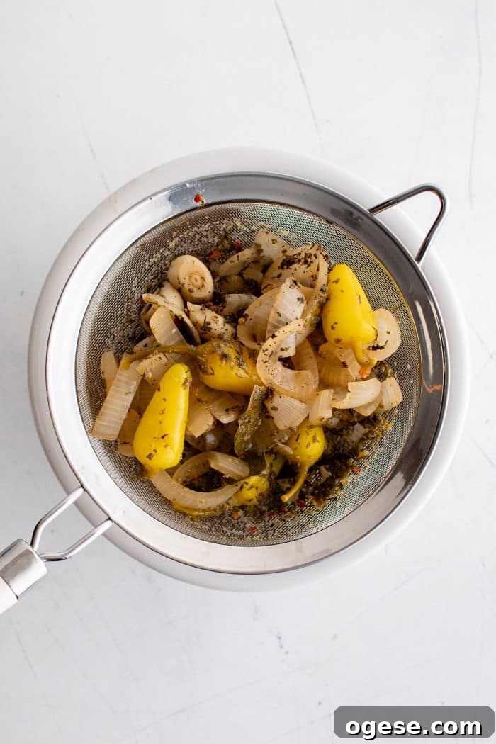 Classic Italian Beef Sandwich Recipe: Tender Slow-Cooked Roast Beef on Italian Bread 10 Fine mesh strainer resting on top of a large white bowl filled with sliced onions, whole pepperoncinis, seasonings, and whole garlic cloves.