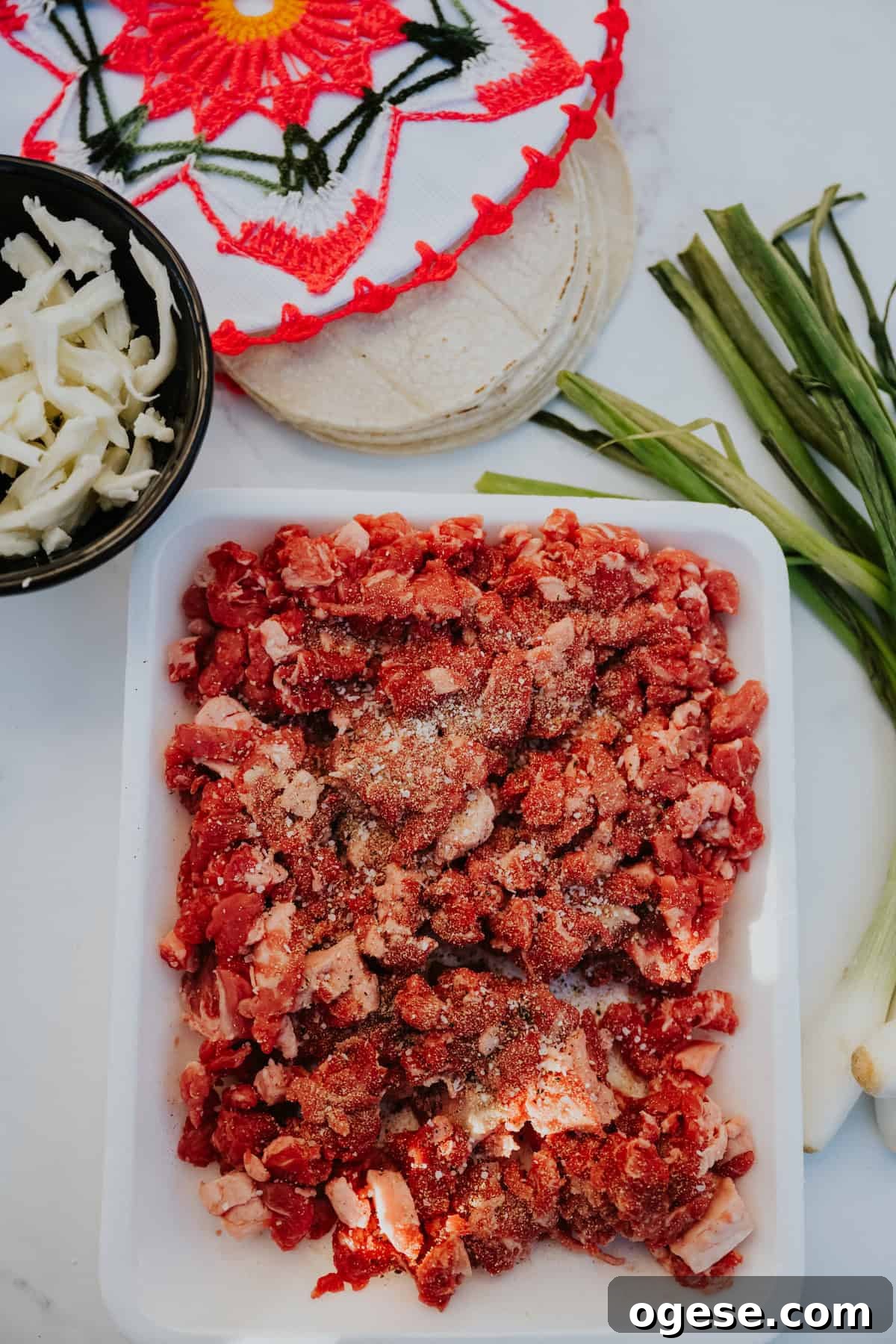 Fresh diced beef taco meat on a white plate surrounded with green onions, corn tortillas, oaxaca cheese.