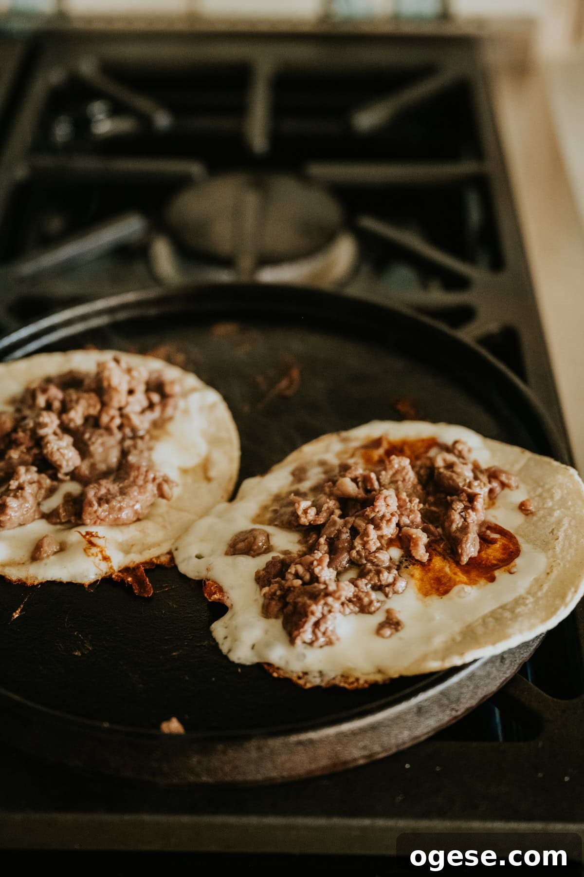 Tacos cooking on a comal with melted cheese and carne asada filling.