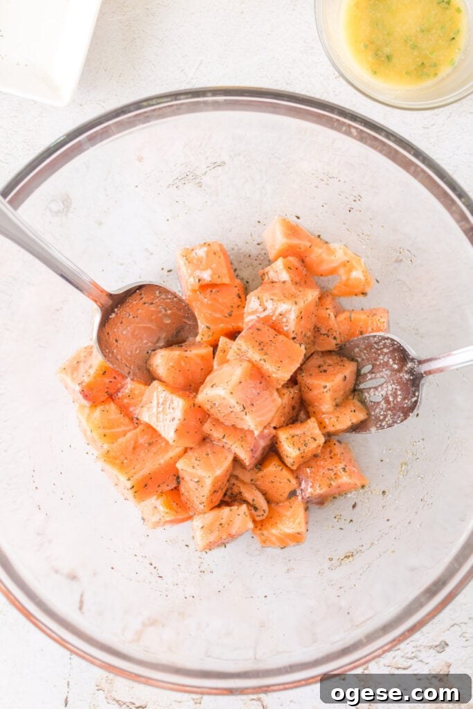Cubes of raw salmon in a glass bowl being mixed with oil and seasonings using two metal spoons.