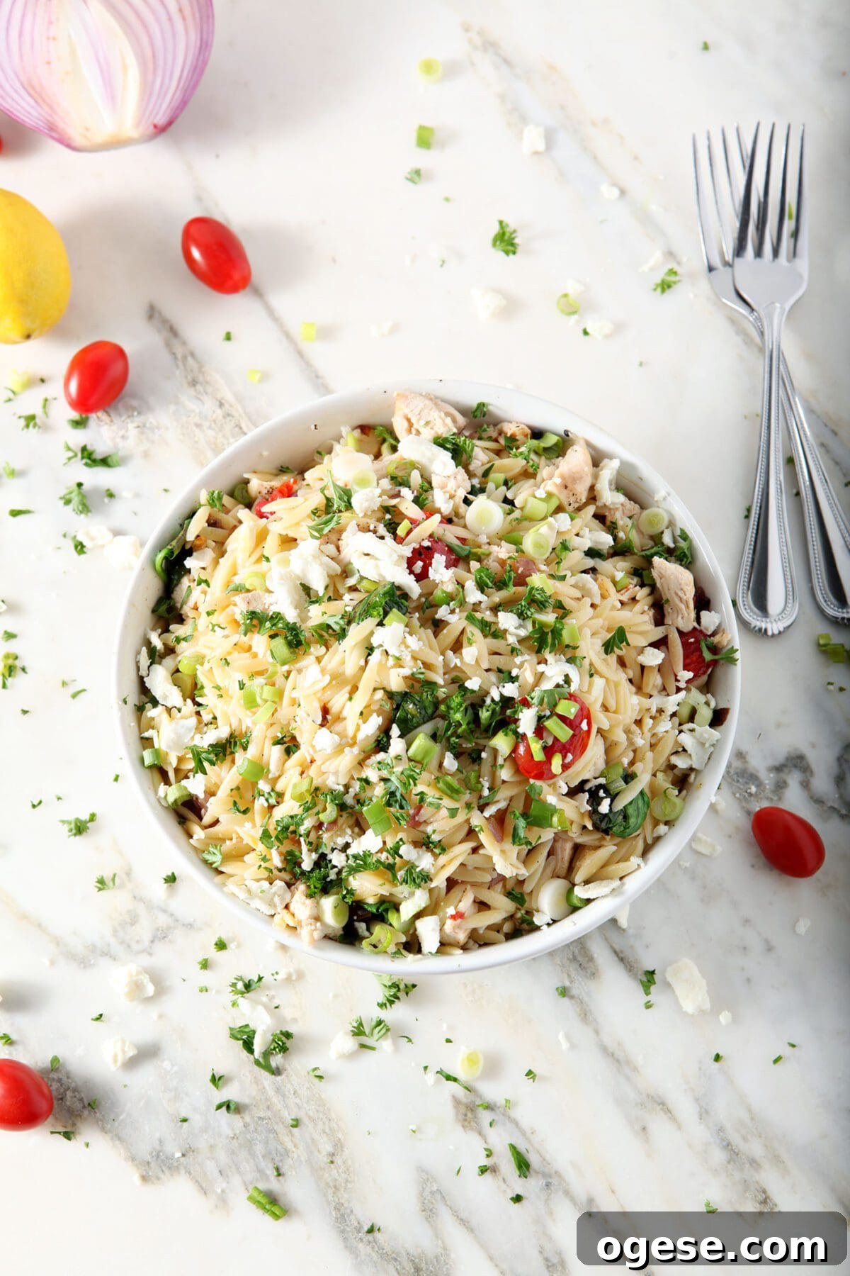 Overhead of a bowl of Greek Orzo Pasta Salad on marble, surrounded by tomatoes, chopped parsley, lemon and red onion