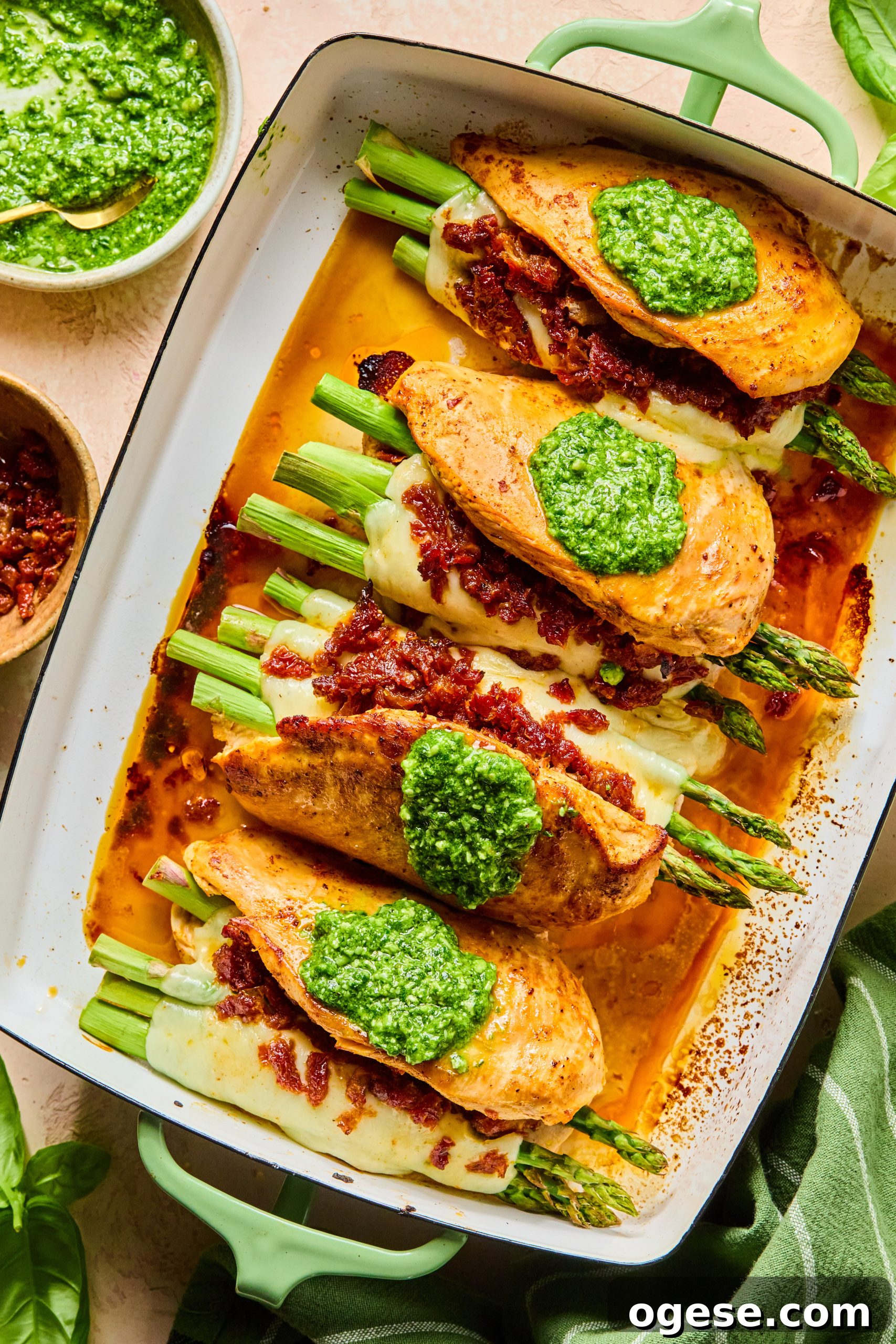 A large baking dish filled with cheesy asparagus stuffed chicken breasts each topped with a spoonful of homemade pesto